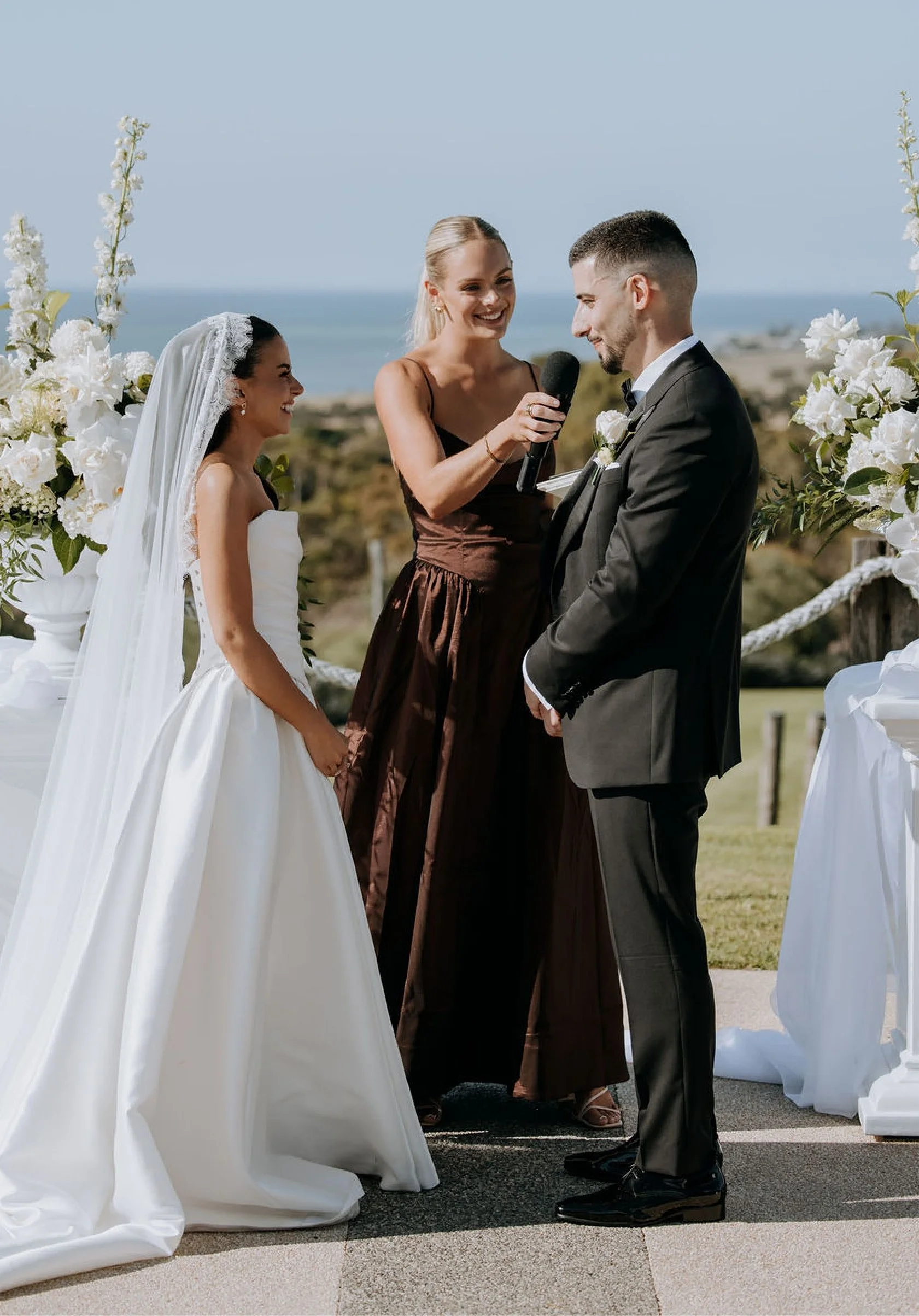 A bride and groom standing outdoors during their wedding ceremony, holding hands and smiling at each other. An officiant is reading vows into a microphone. The scene is decorated with white flowers and greenery, with a scenic view of the ocean and sky in the background.