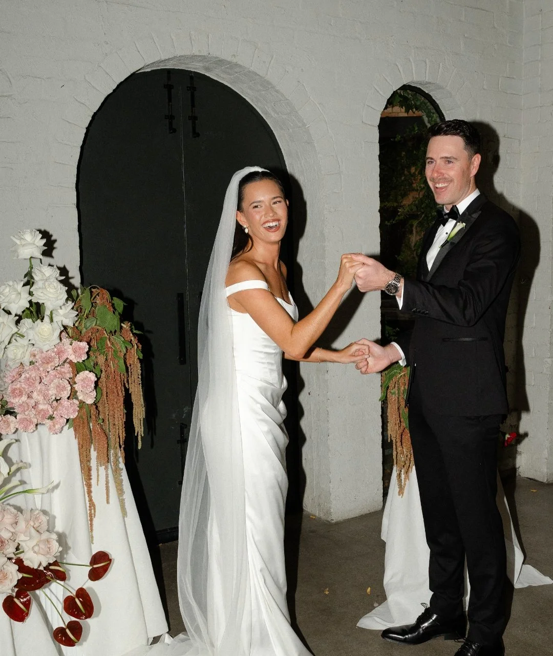 Bride and groom holding hands and smiling at each other during wedding ceremony in front of floral arrangements.