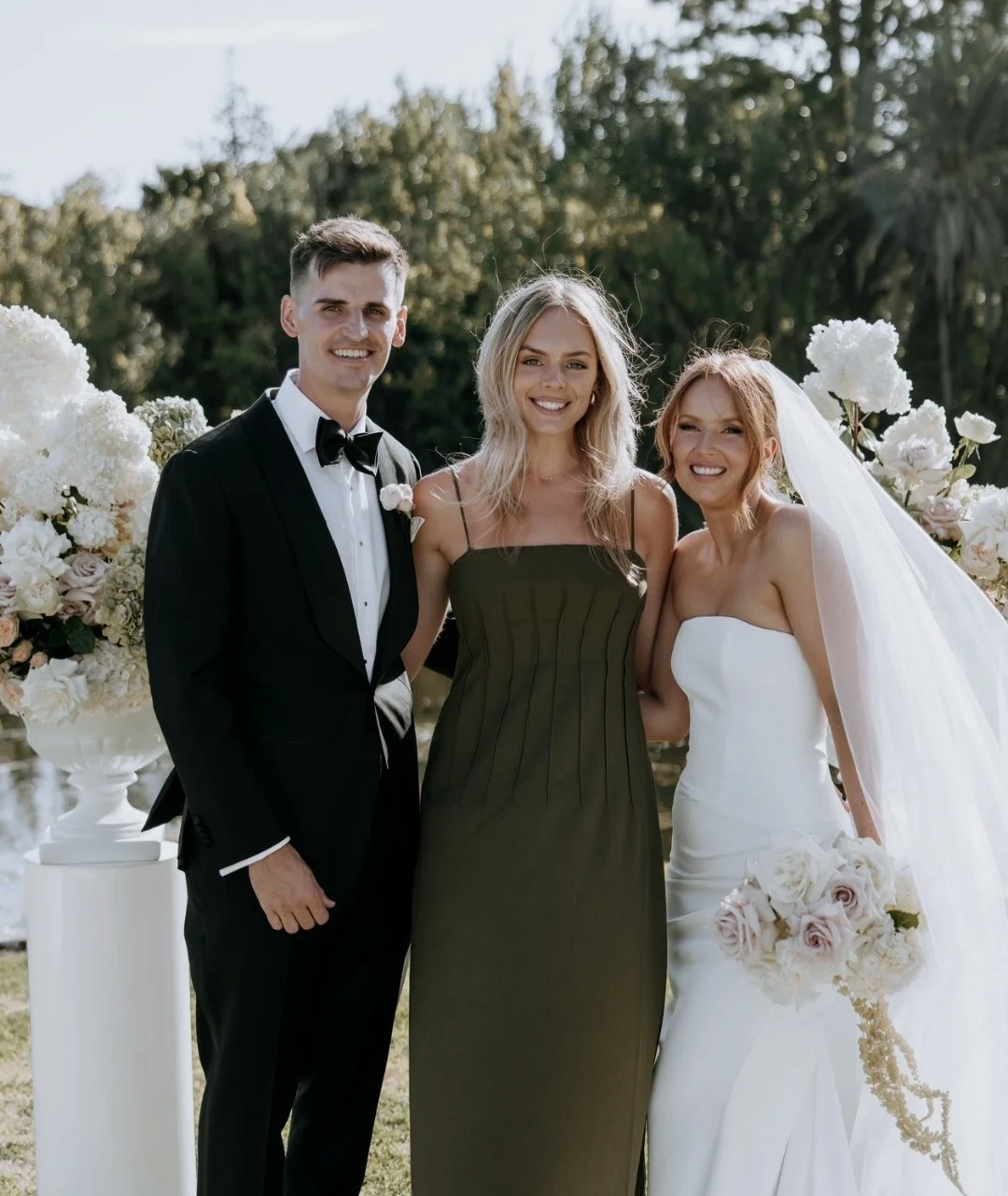 Group of three people dressed in formal wedding attire standing outdoors with floral arrangements in the background.