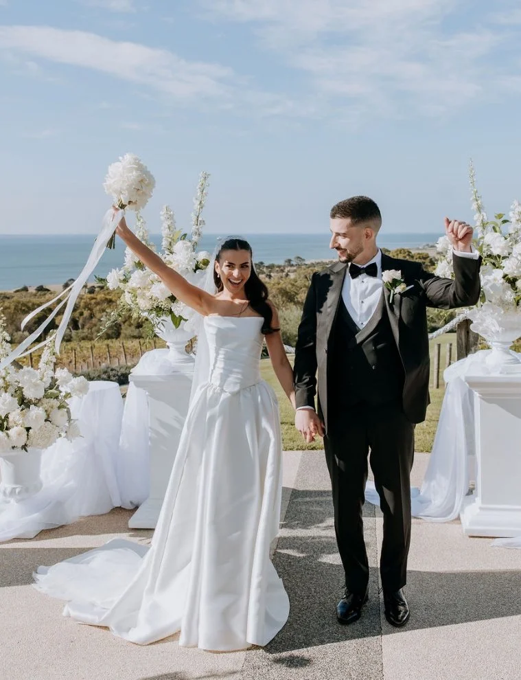 A newlywed couple celebrating outdoors with a scenic view of the ocean in the background. The bride is holding a bouquet and wearing a white wedding gown, and the groom is dressed in a black tuxedo with a bow tie. They are smiling and holding hands amid an arrangement of white floral decorations.