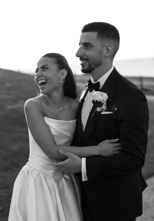 A happy bride and groom in wedding attire sharing a joyful moment outdoors.