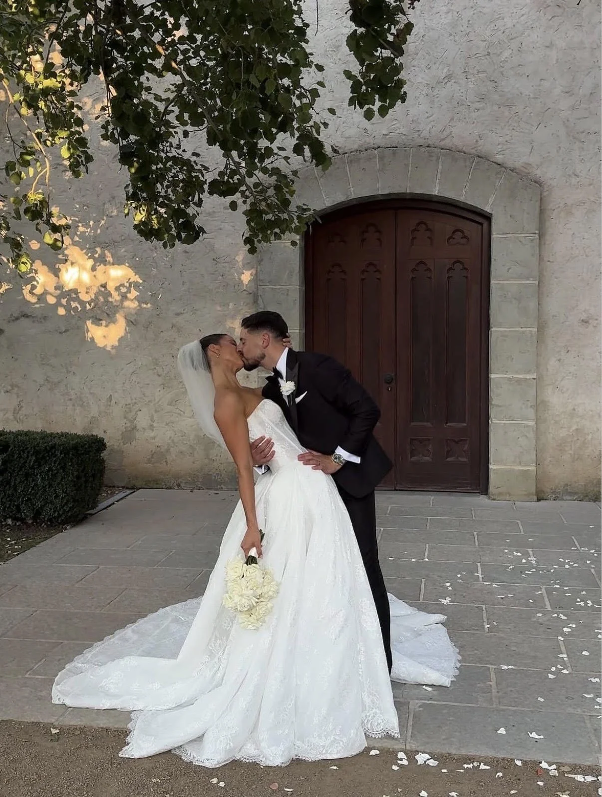 A bride and groom kissing outdoors in front of a stone building with a wooden door. The bride is in a white strapless wedding gown holding a bouquet of white flowers, and the groom is in a black suit with a white shirt and black tie. They are leaning into each other, with scattered flower petals on the ground.
