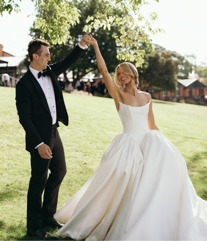 A newlywed couple dancing outdoors on a sunny day, with the groom wearing a black tuxedo and the bride in a white wedding gown, smiling and twirling.