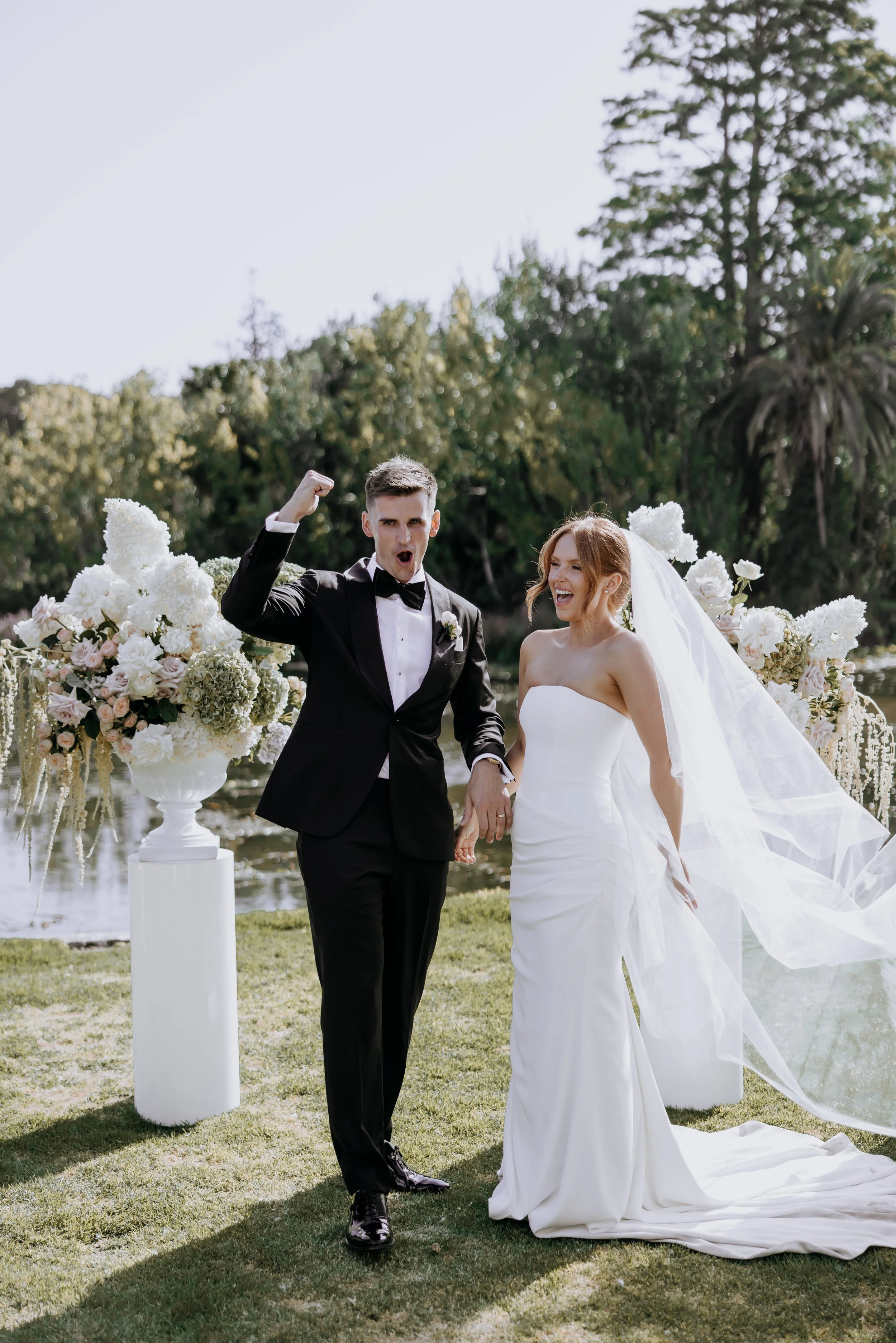 A newlywed couple celebrating outdoors by a lake, with the groom in a tuxedo and the bride in a strapless wedding gown, surrounded by floral arrangements, trees, and a bright sky.
