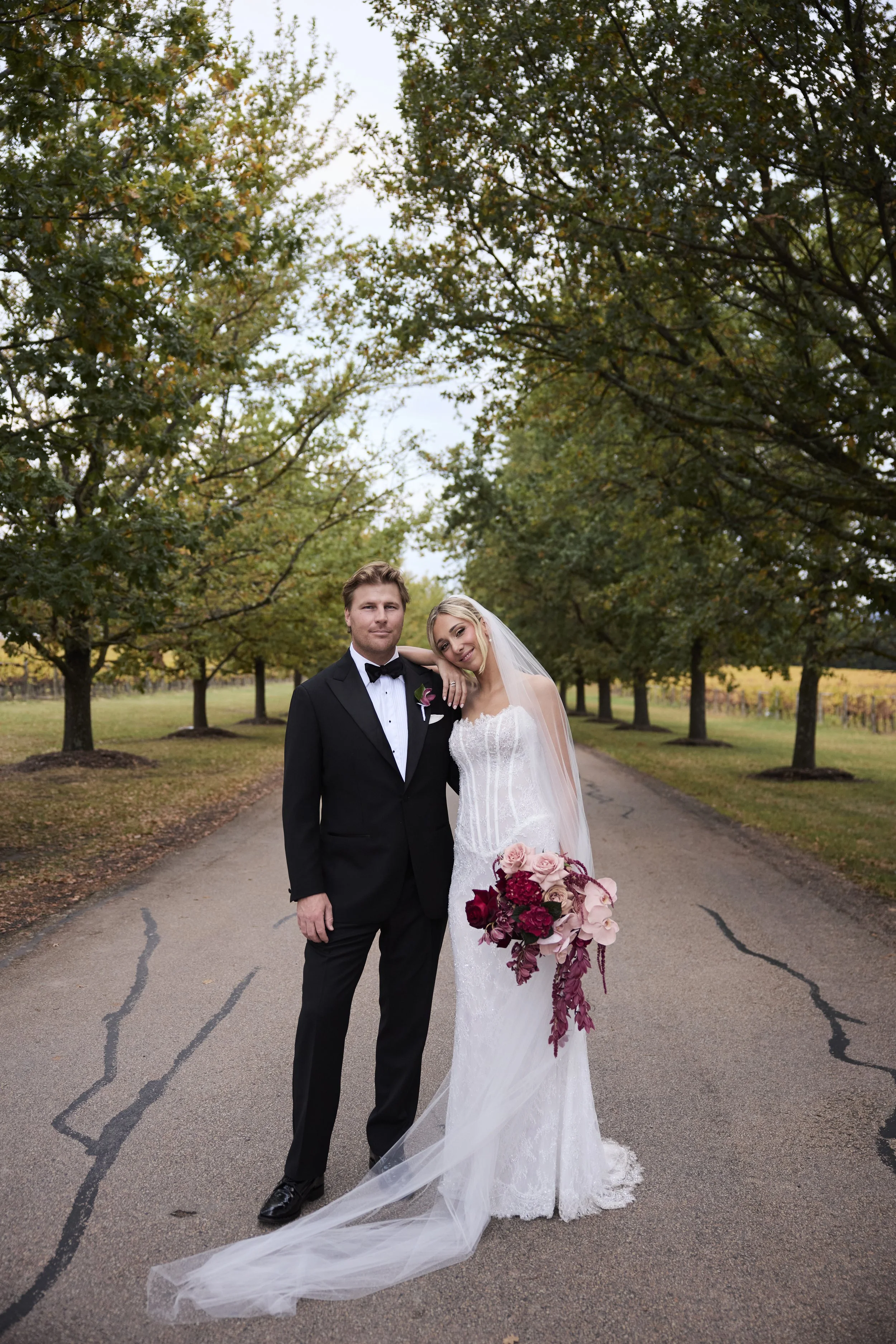 A bride and groom standing together on a tree-lined road during daytime, dressed in wedding attire, with the bride holding a large bouquet of pink, red, and burgundy flowers, and the groom wearing a black tuxedo and bow tie.