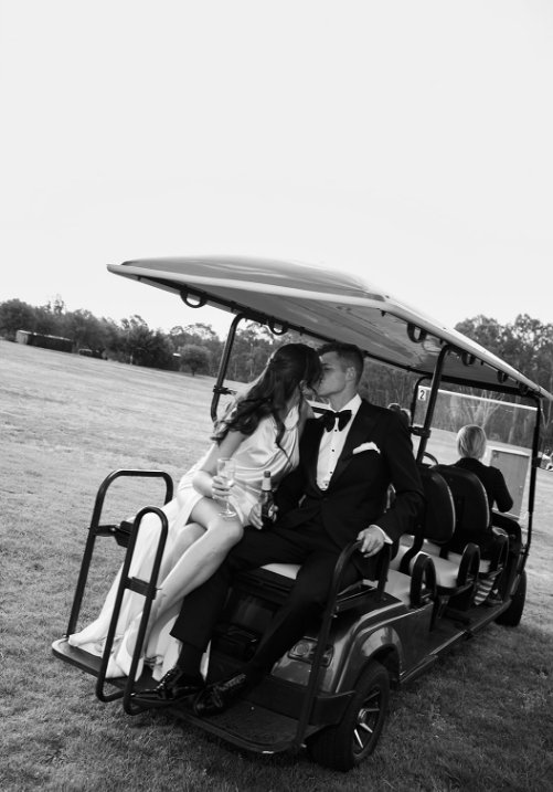 A couple in formal attire sitting on the front seat of a golf cart, sharing a kiss, with a woman in a white dress and a man in a tuxedo. In the background, two other people are seated in the golf cart, and an open field with trees is visible.