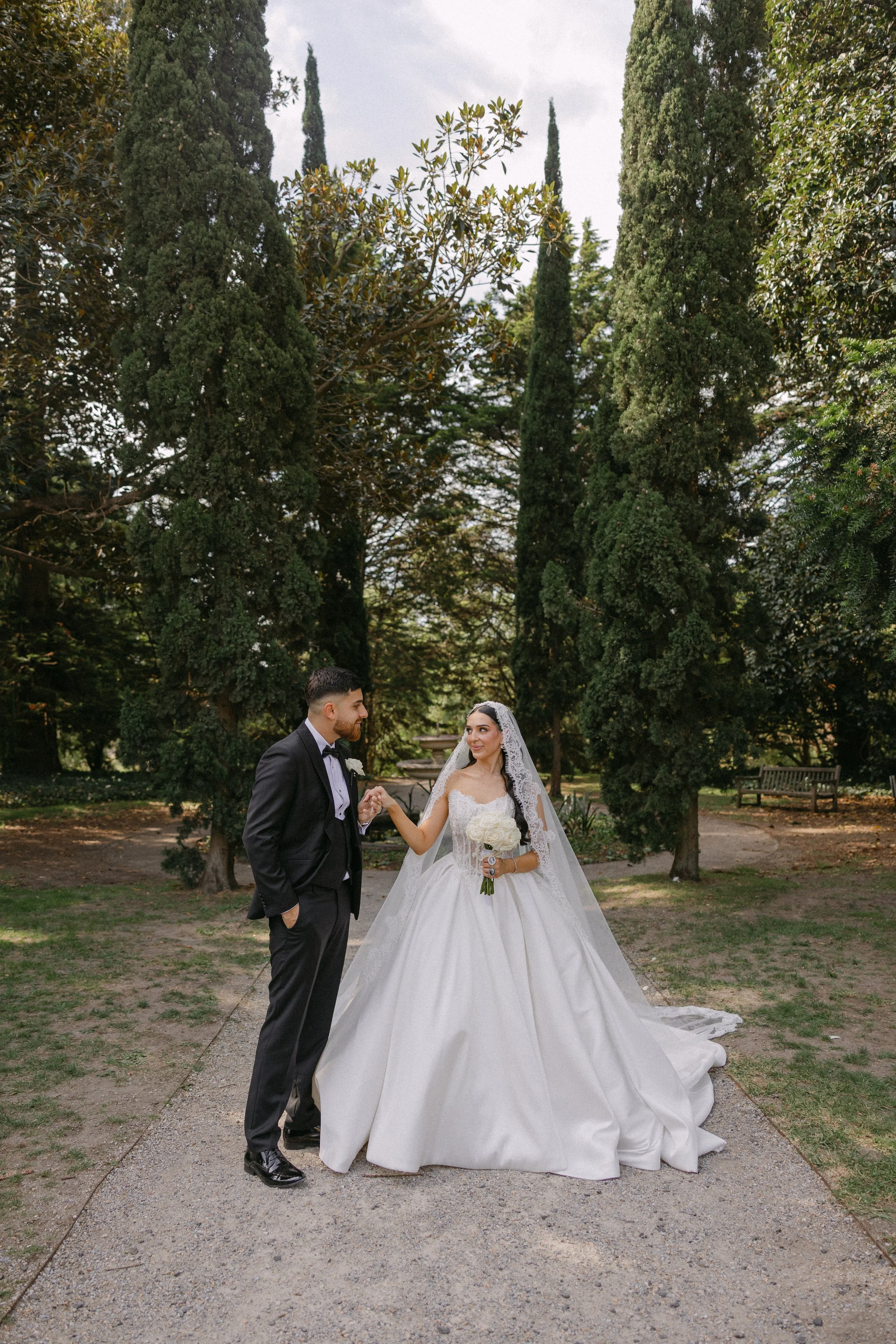 A bride and groom hold hands and look at each other in a park with tall trees and benches in the background.