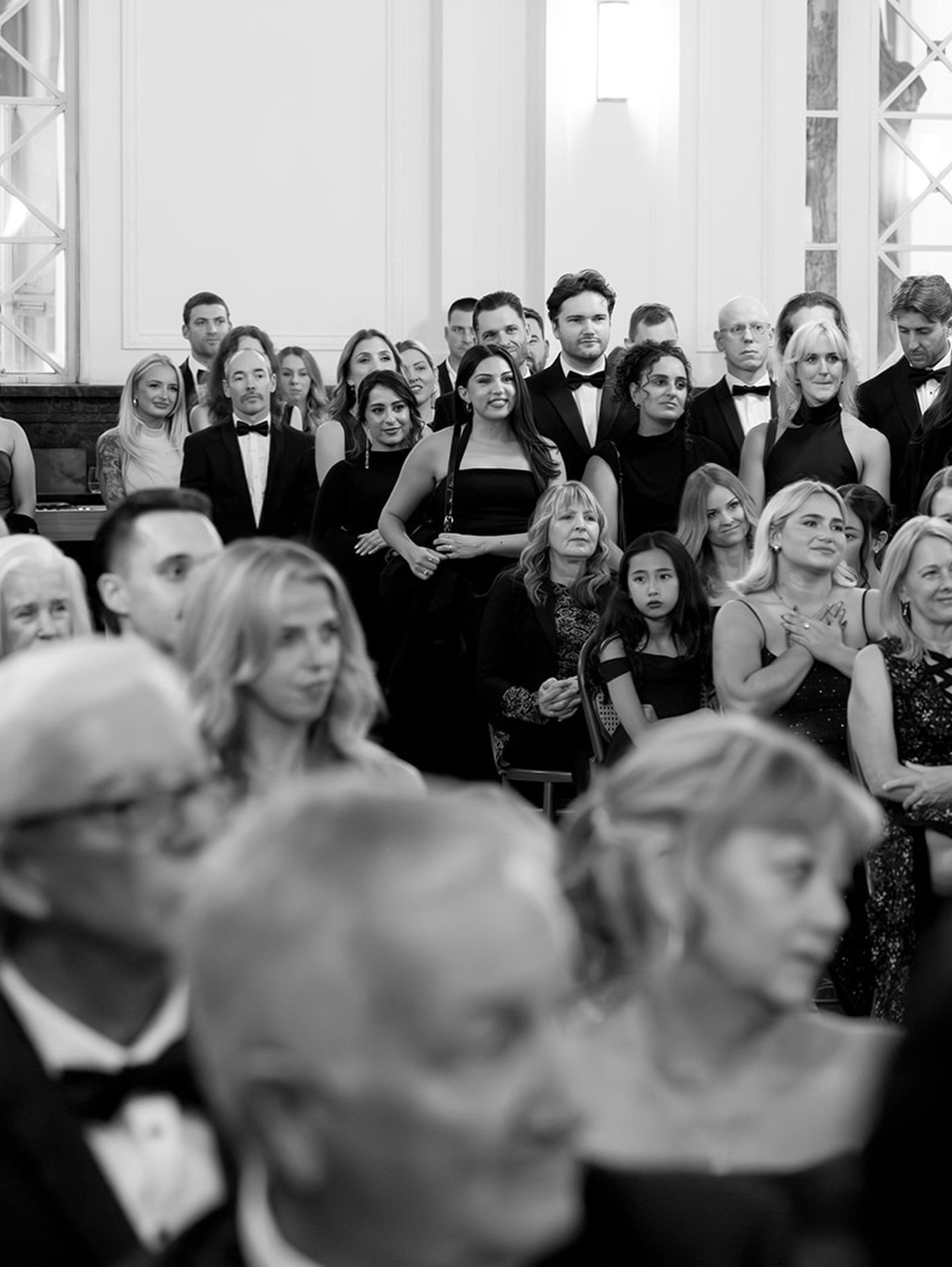 Black and white photo of a group of people dressed formally, standing closely together indoors, some smiling, facing forward, with a woman in the center wearing a sleeveless dress.
