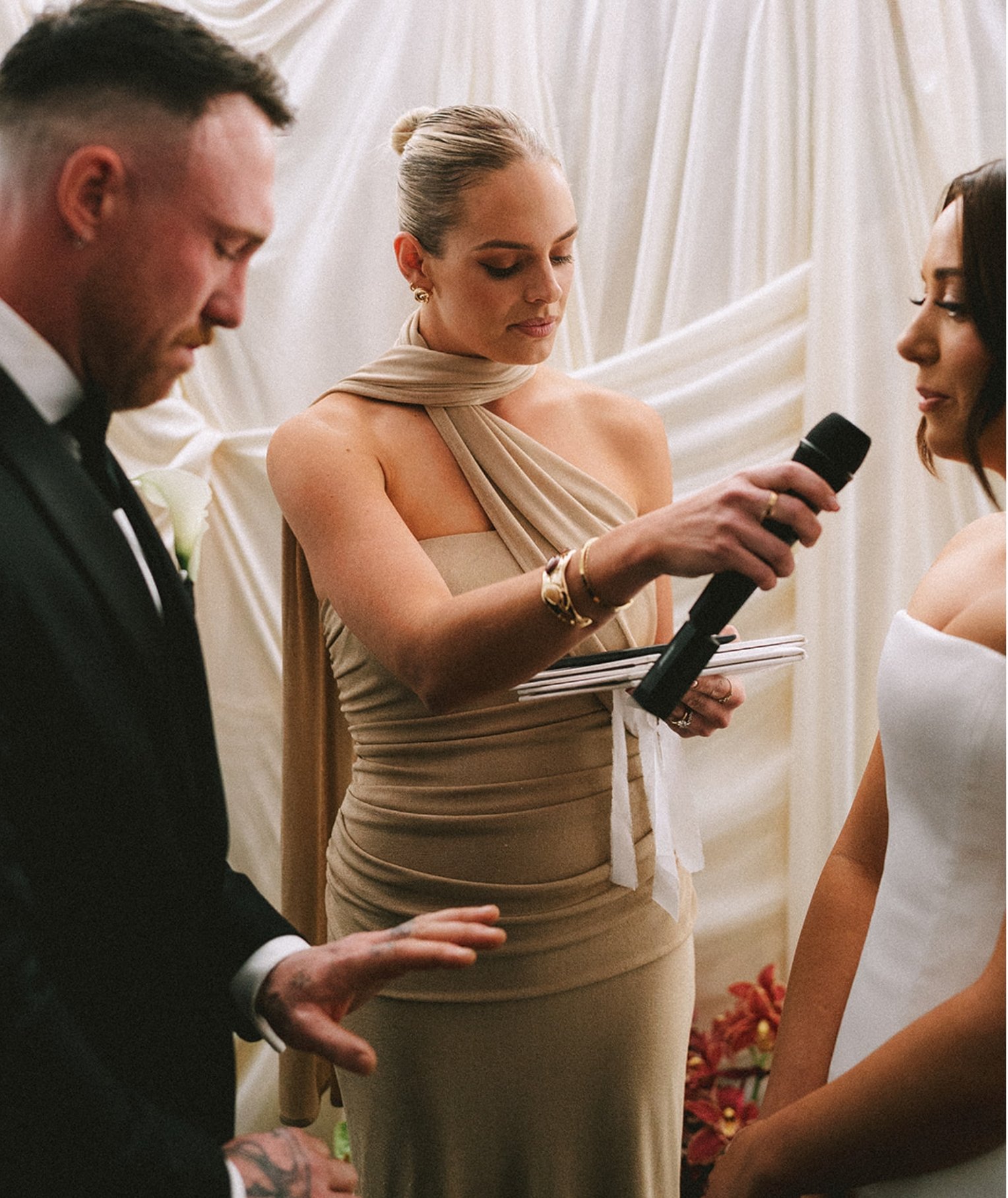 A wedding ceremony with a couple holding hands, a woman reading vows into a microphone, and an officiant standing between them, all in formal attire with a white draped backdrop.