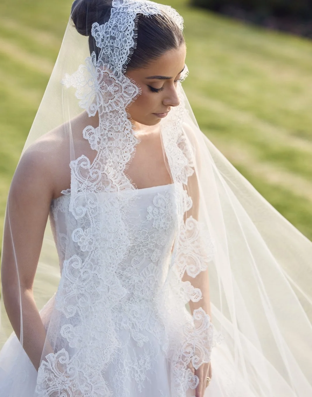 A bride in a white lace wedding gown and a veil with lace details, standing outdoors on a grassy area, with her head slightly bowed.