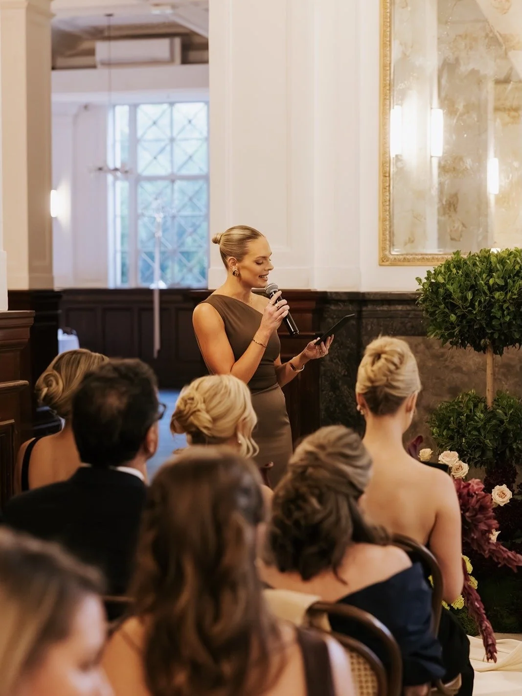 A woman in a brown dress is giving a speech or toast at a formal event, possibly a wedding, while holding a microphone and reading from a tablet. There are several seated guests, mostly women, in elegant attire, with a decorative indoor setting inclu