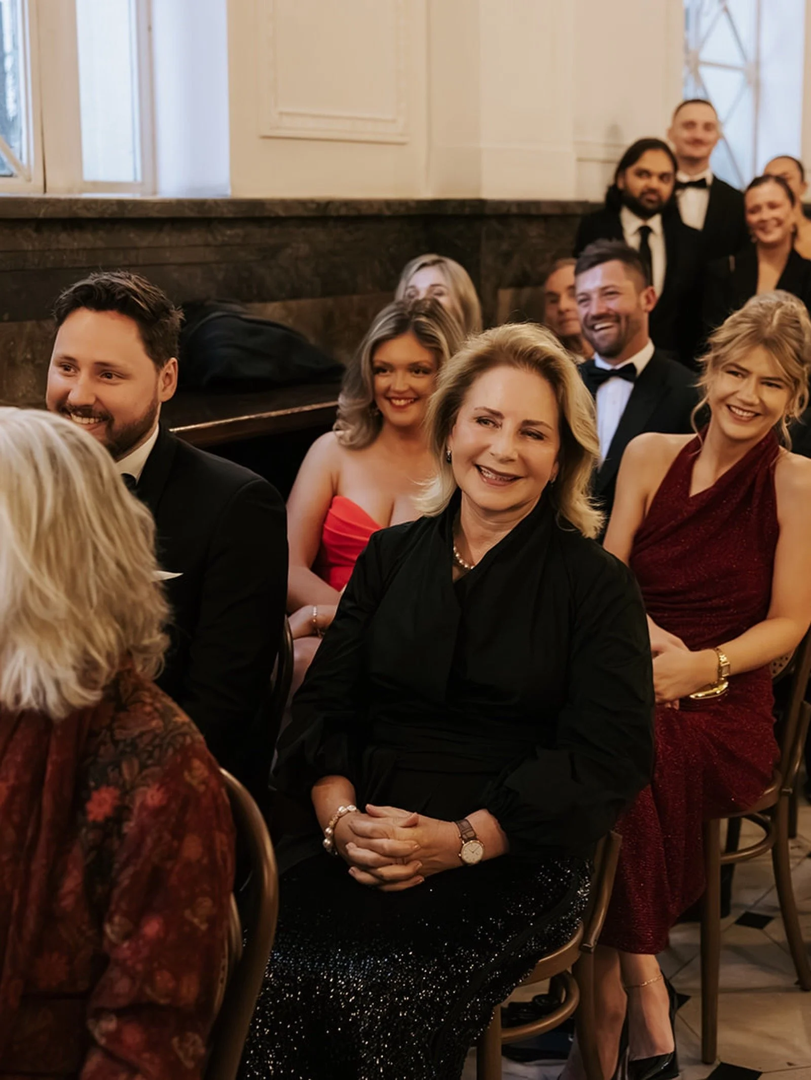 A group of formally dressed people at a social event, sitting and smiling, including women in elegant dresses and men in tuxedos, in a room with high windows and marble walls.