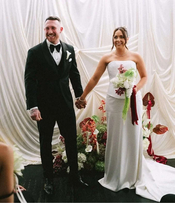A bride and groom holding hands during their wedding ceremony, standing in front of a decorative draped backdrop with floral arrangements.