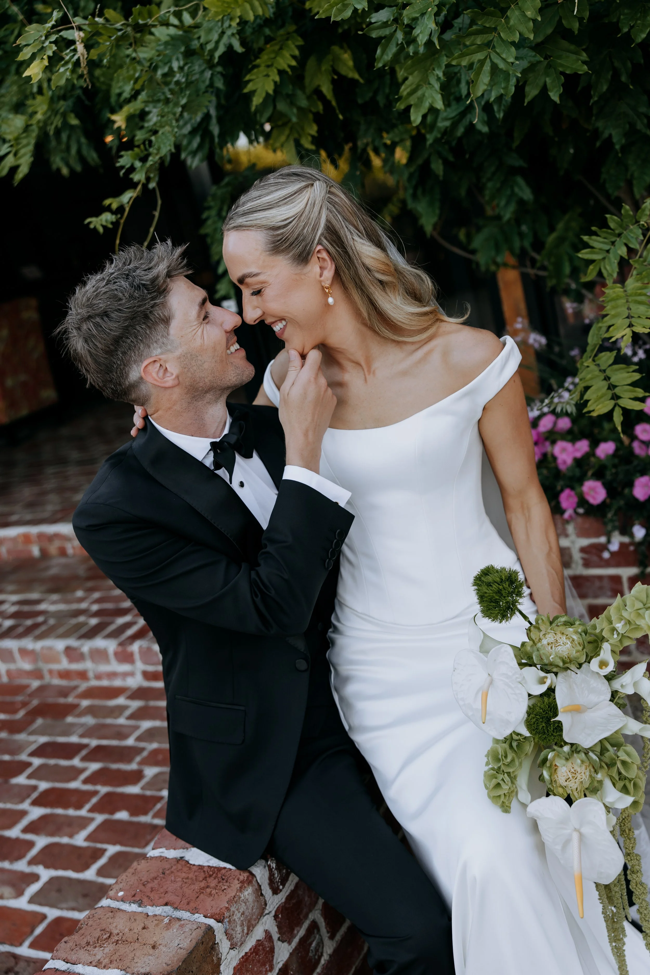 A newlywed couple in elegant wedding attire sharing a tender moment outdoors, with the groom in a black tuxedo and the bride in a white off-shoulder wedding gown, surrounded by lush greenery and colorful flowers.