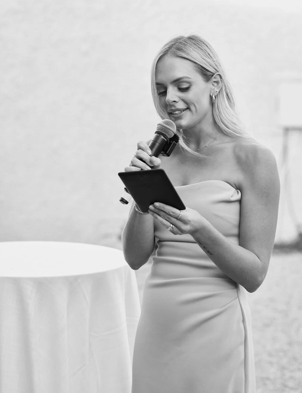 A woman in a strapless dress holding a microphone and reading from a tablet.