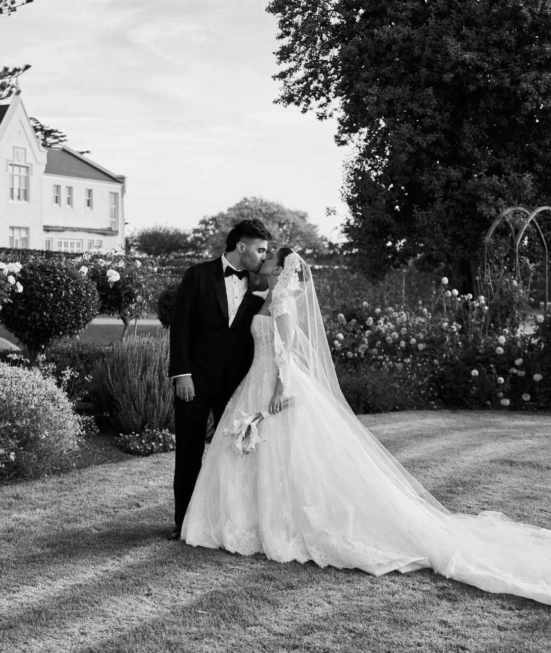 A bride and groom sharing a kiss outdoors, with the groom in a black tuxedo and the bride in a white wedding gown and veil, holding a bouquet of flowers in a garden setting.