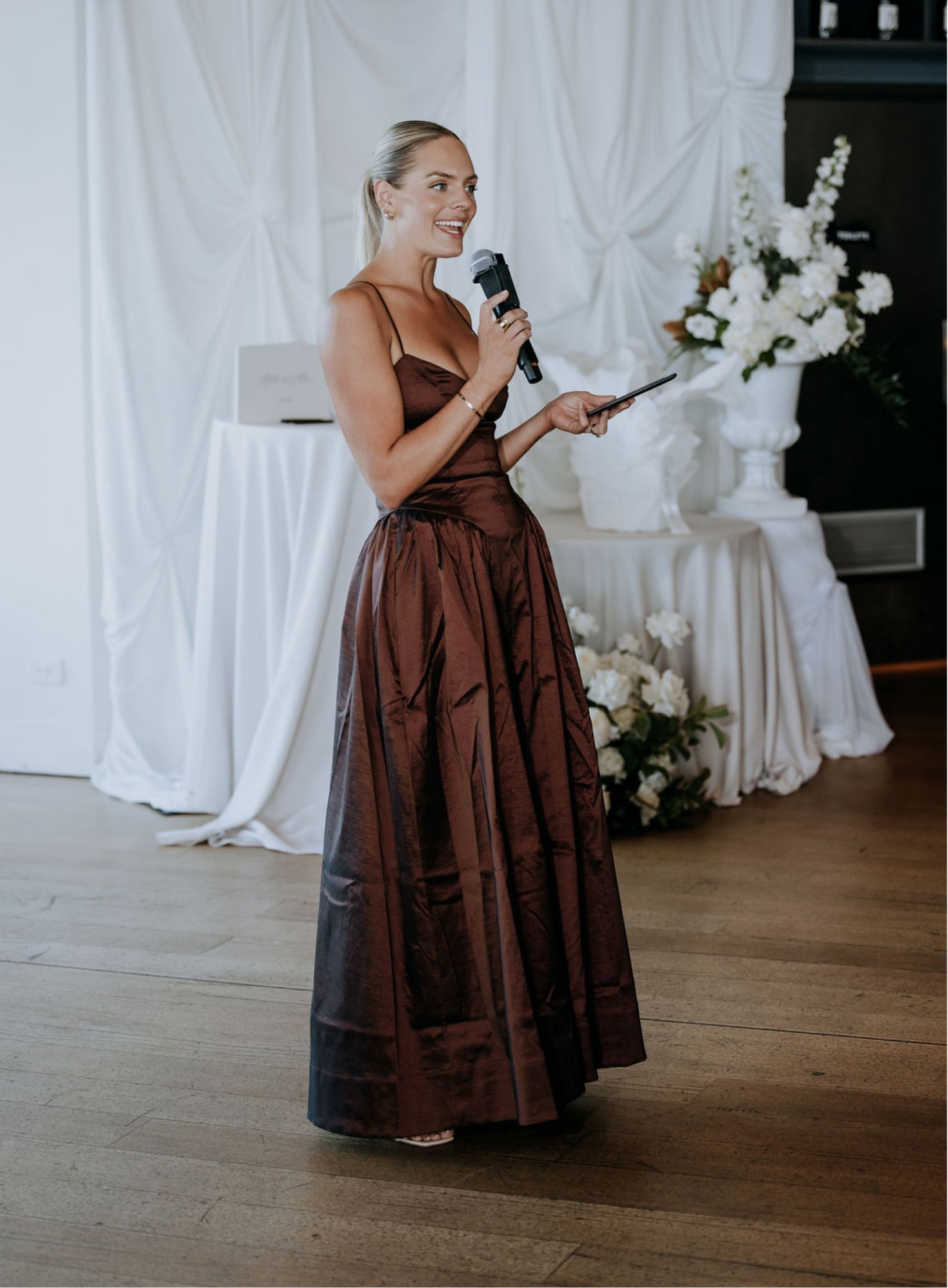 A woman in a long, brown dress, holding a microphone and a tablet, standing in front of a backdrop with white fabric and floral arrangements at an indoor event.