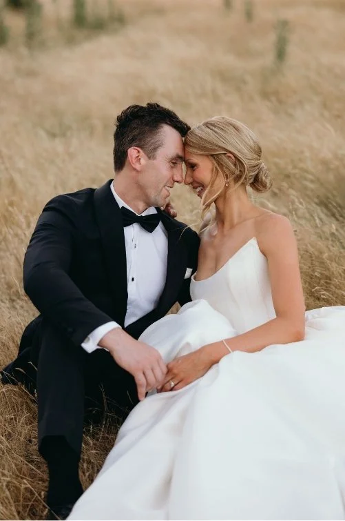 A bride and groom sitting close together in a field, touching foreheads and smiling at each other.