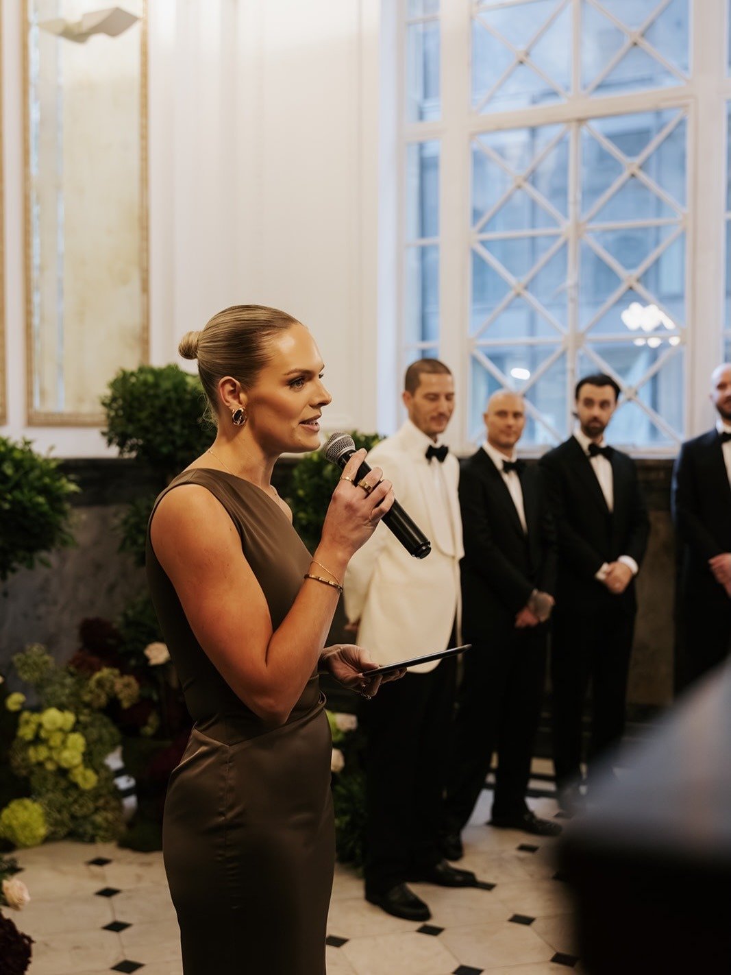 A woman in a brown dress speaking into a microphone at what appears to be a formal event, with men in tuxedos standing in the background.