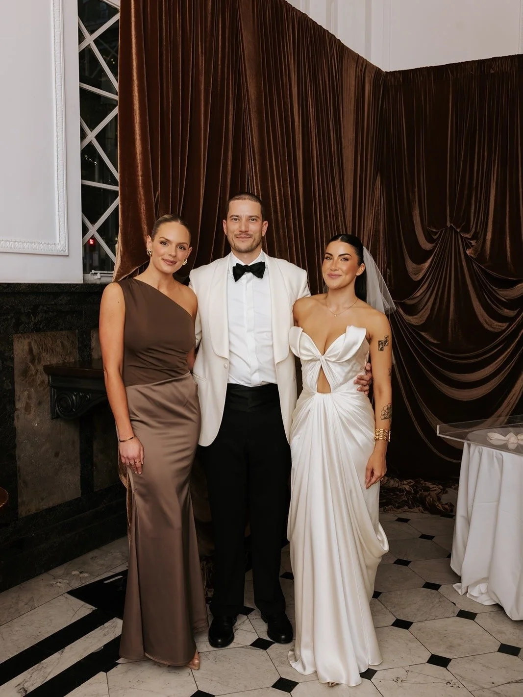 Three people dressed in formal attire at a special event. The person in the middle is wearing a white tuxedo with a black bow tie, flanked by two women in elegant gowns, one in brown and one in white.