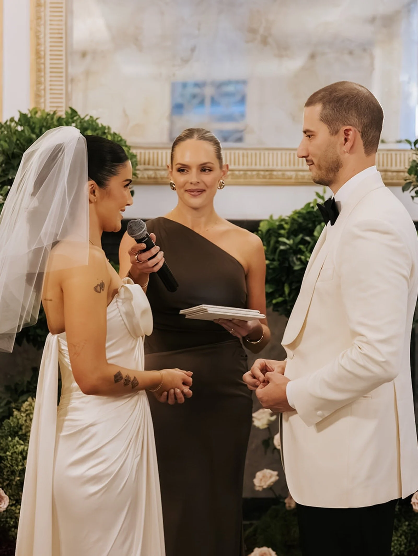 A bride and groom exchanging vows during a wedding ceremony, with a woman officiant standing between them holding a microphone and a notepad.