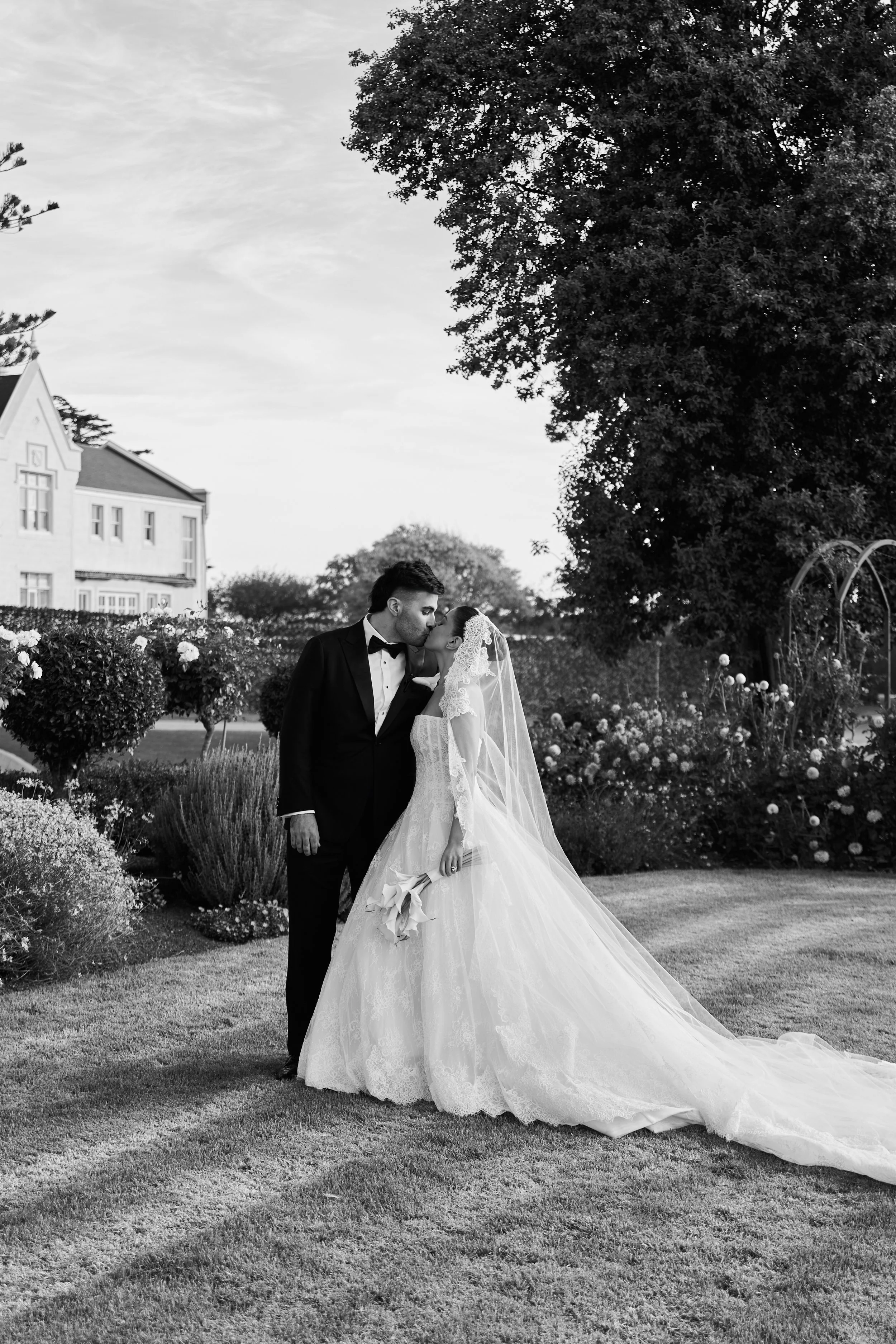 A black and white photo of a bride and groom kissing outdoors in a garden, with trees and flowers in the background.