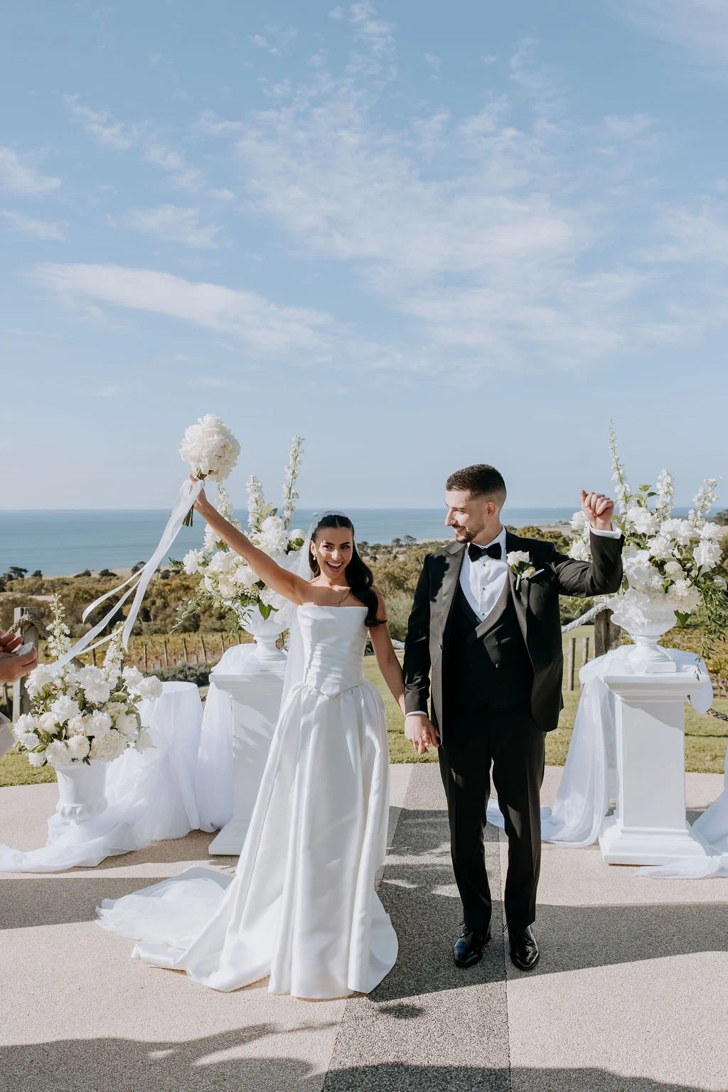 A bride and groom holding hands, celebrating outdoors with flowers and scenic ocean view in the background