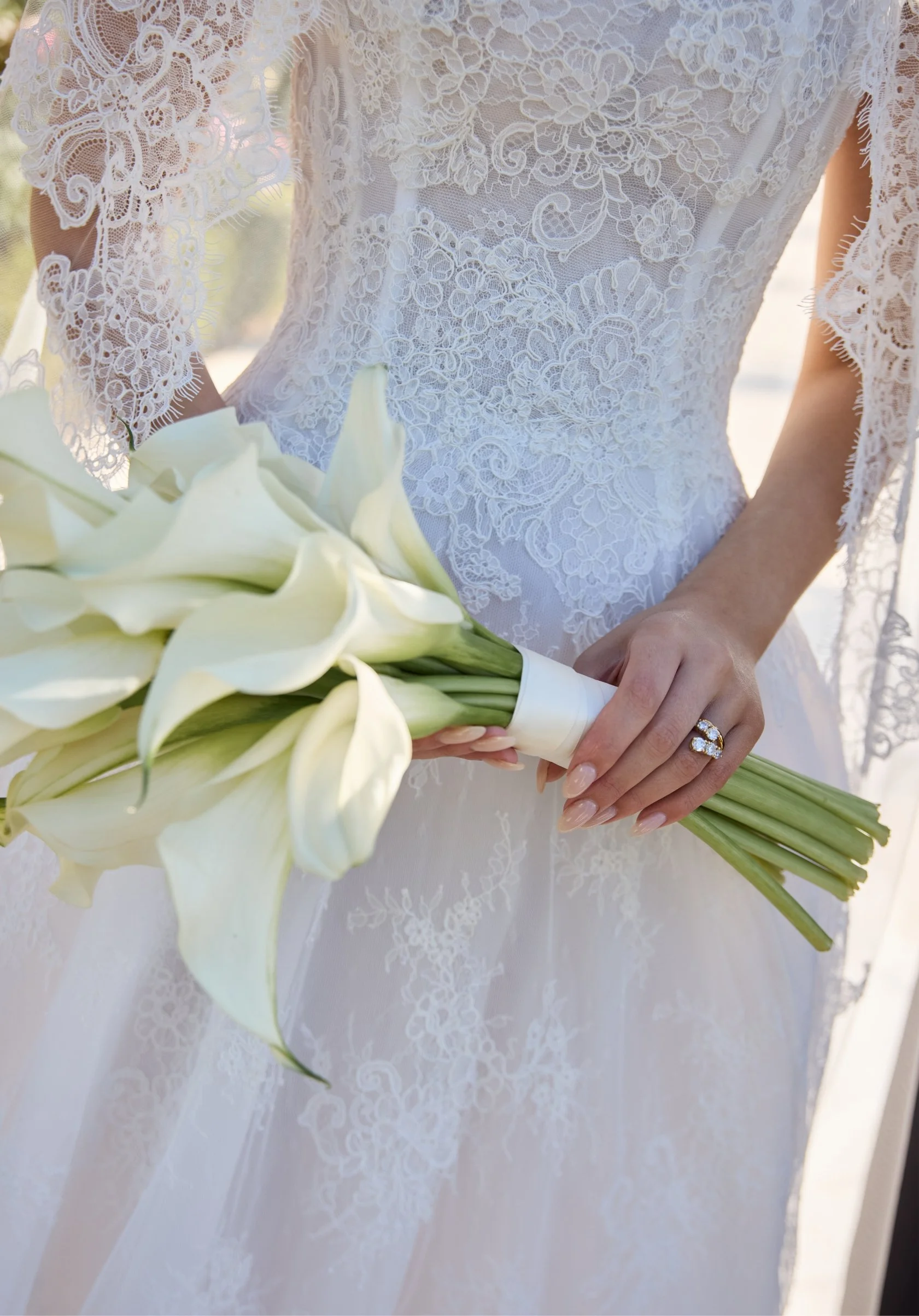 Close-up of a bride in a detailed lace wedding gown holding a bouquet of white calla lilies.