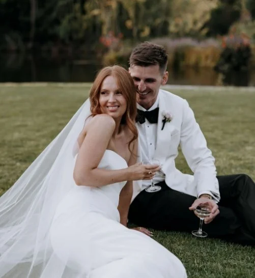 A bride and groom sitting on grass by a lake, smiling and holding glasses of champagne during their outdoor wedding.