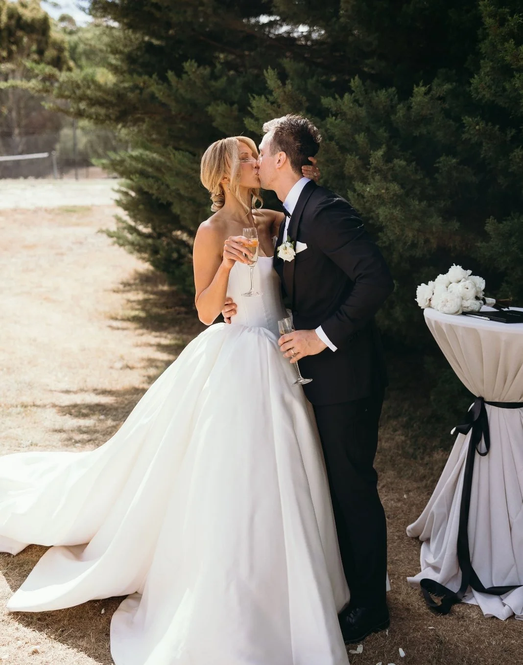 Bride and groom sharing a kiss outdoors during their wedding, holding champagne glasses, with wedding decor nearby.