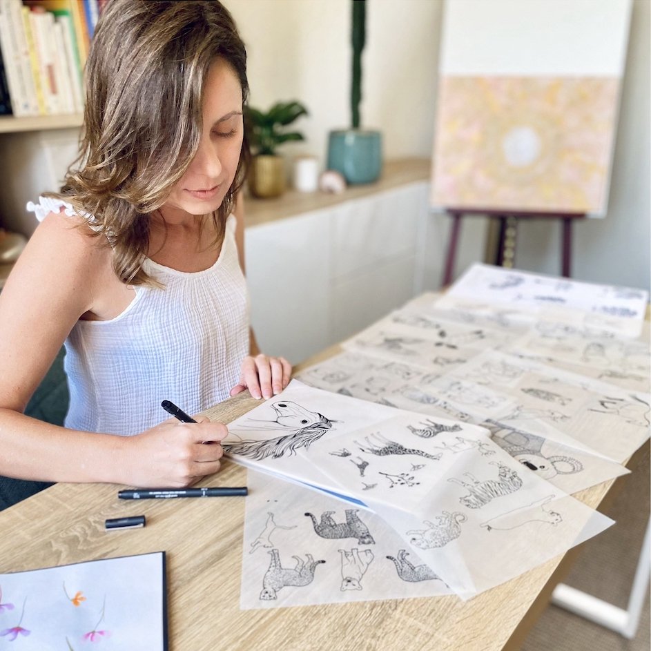 Lady siting by a desk drawing illustration of animals in studio with books, plants and painting in the background