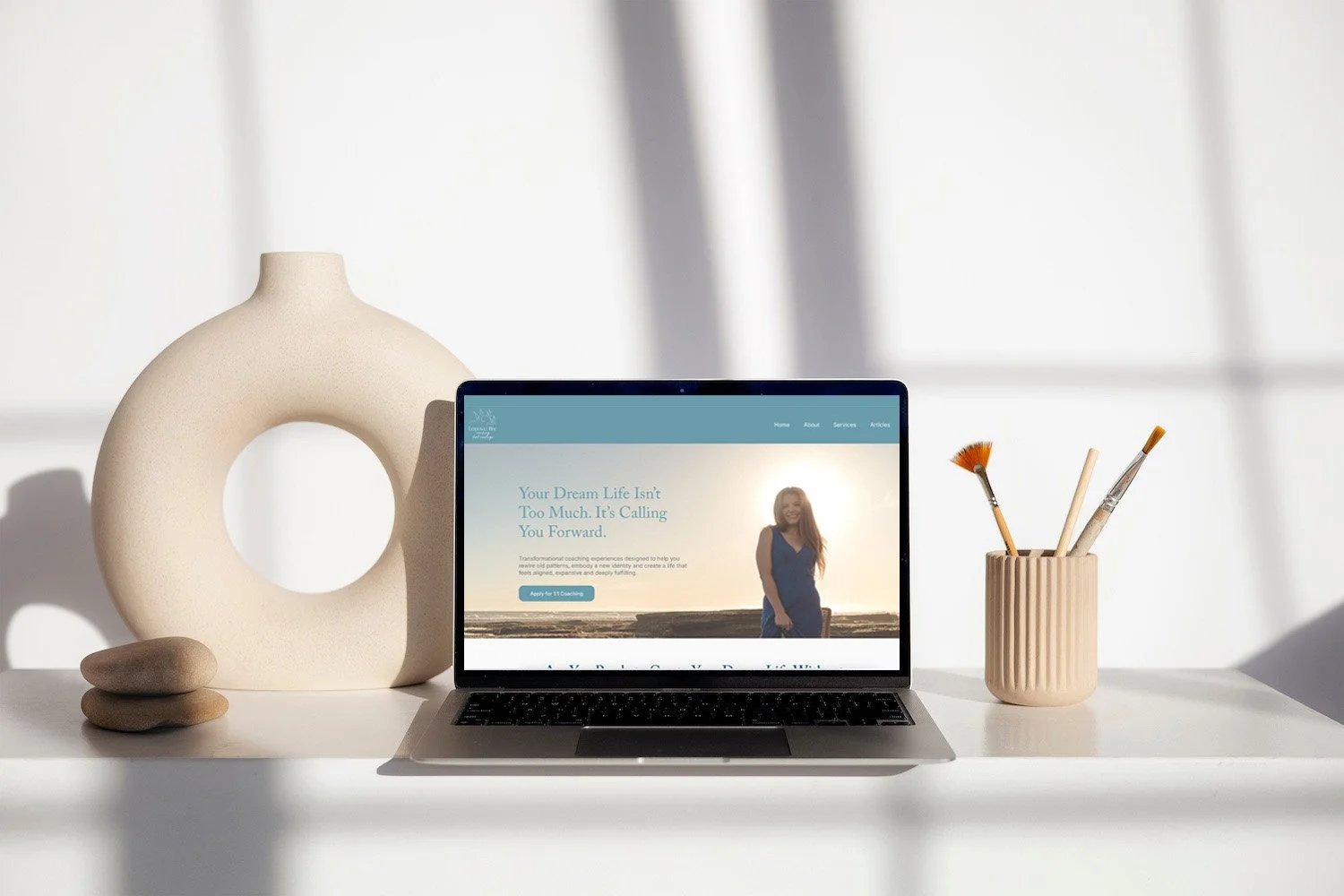 A laptop on a white desk displaying a website with a woman on the screen, surrounded by a decorative vase, rocks, and a cup holding paintbrushes in a minimalist setting with a bright window in the background.