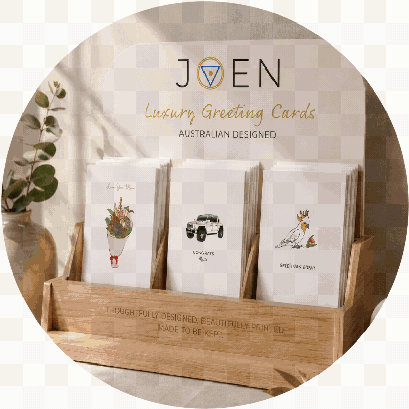 Australian Display of luxury greeting cards in a wooden holder at a store, with a potted plant to the side and a sign above. The cards feature illustrations and messages. Melbourne, Geelong