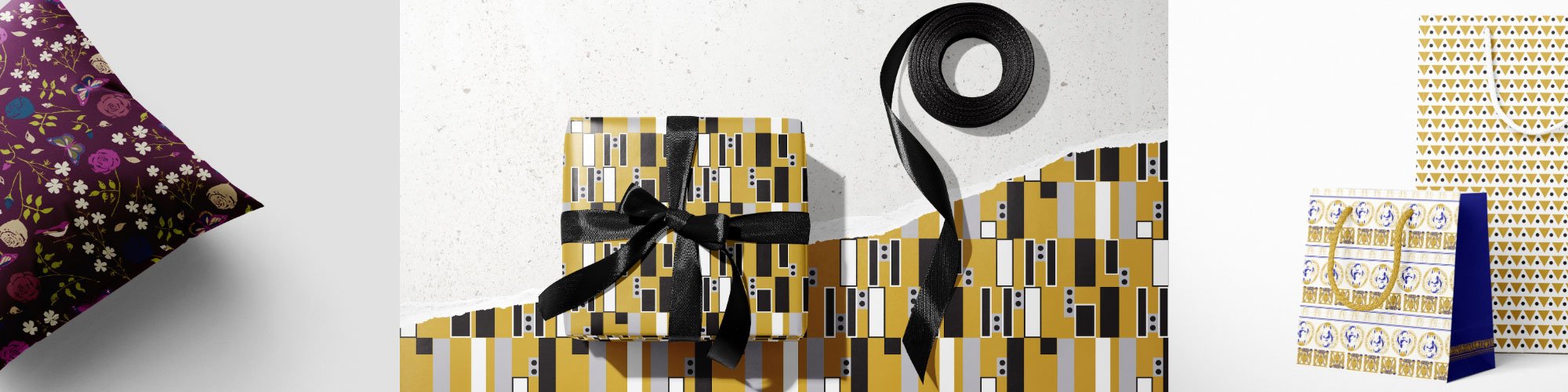 Decorative gift wrapped in black and gold geometric patterned paper with a black ribbon, alongside a roll of black ribbon on a white and patterned background. Wrapping paper.