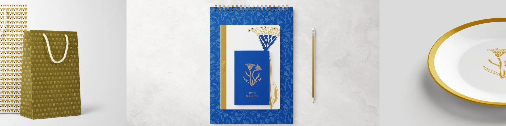 A collection of gold and white patterned gift bags, a blue and white floral-themed planner with a gold spine, and a white ceramic plate with a gold rim and floral design, displayed on a light-colored surface. Stationary.