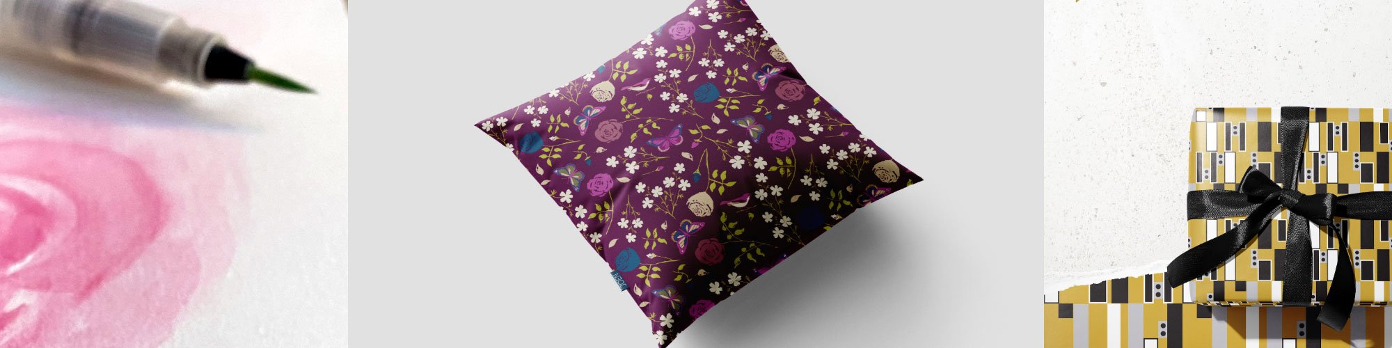 A decorated gift box with geometric patterns and a black ribbon, a floral-patterned pillow, and a piece of pink watercolor alongside a pen. Home decor.