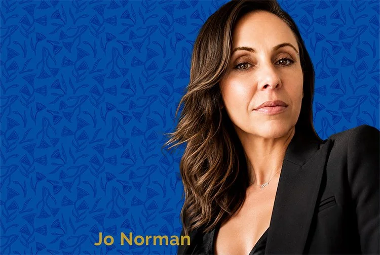 A woman with wavy brown hair wearing a black blazer poses for photo,  a blue background with a subtle pattern. The name Jo Norman appears in yellow text at the bottom left corner.
