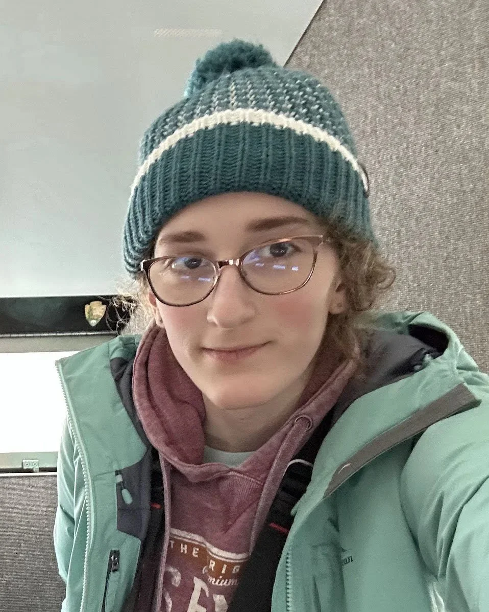 A young woman with glasses, curly hair, wearing a knitted beanie, a maroon hoodie, and a mint green jacket, taking a selfie indoors.