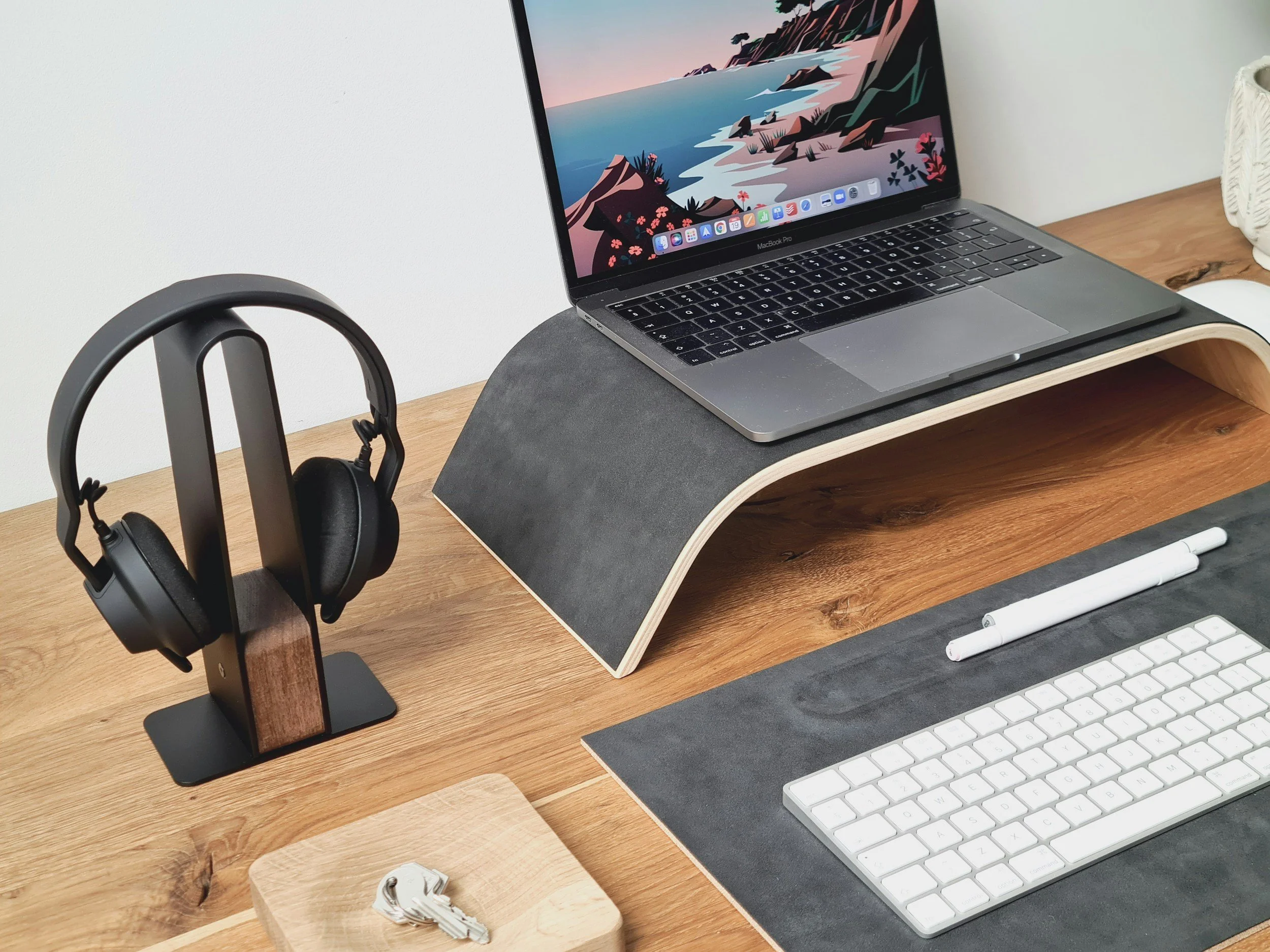 Un bureau en bois avec un ordinateur portable, un casque sans fil sur un support en bois, un clavier blanc, deux stylos blancs, et un support pour écouteurs. L'écran de l'ordinateur affiche un paysage de côte en style d'illustration.