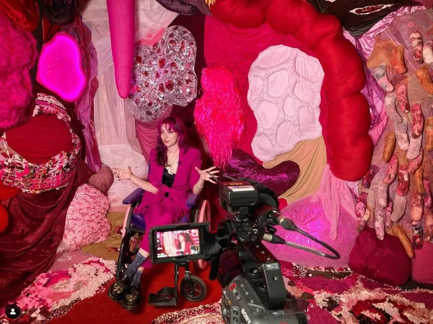 The artist, wearing a pink suit, speaking in front of a video camera within an installation of pink, organic textile forms.