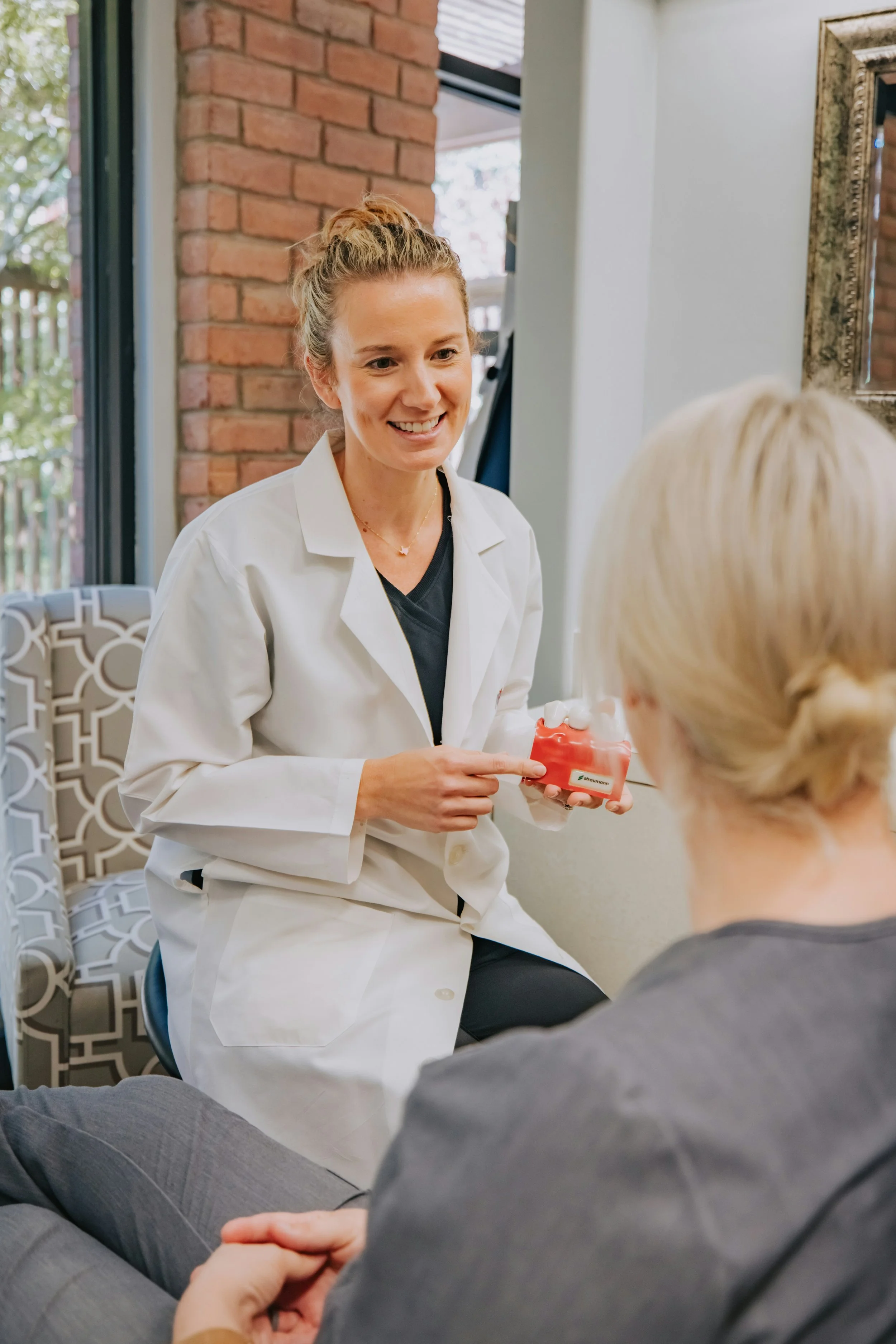 A female healthcare professional, dressed in a white lab coat, sitting and smiling while showing a model of dental gums and teeth to an elderly woman with blonde hair, seated in front of her. The setting appears to be a dental or healthcare office with a brick wall and a window in the background.