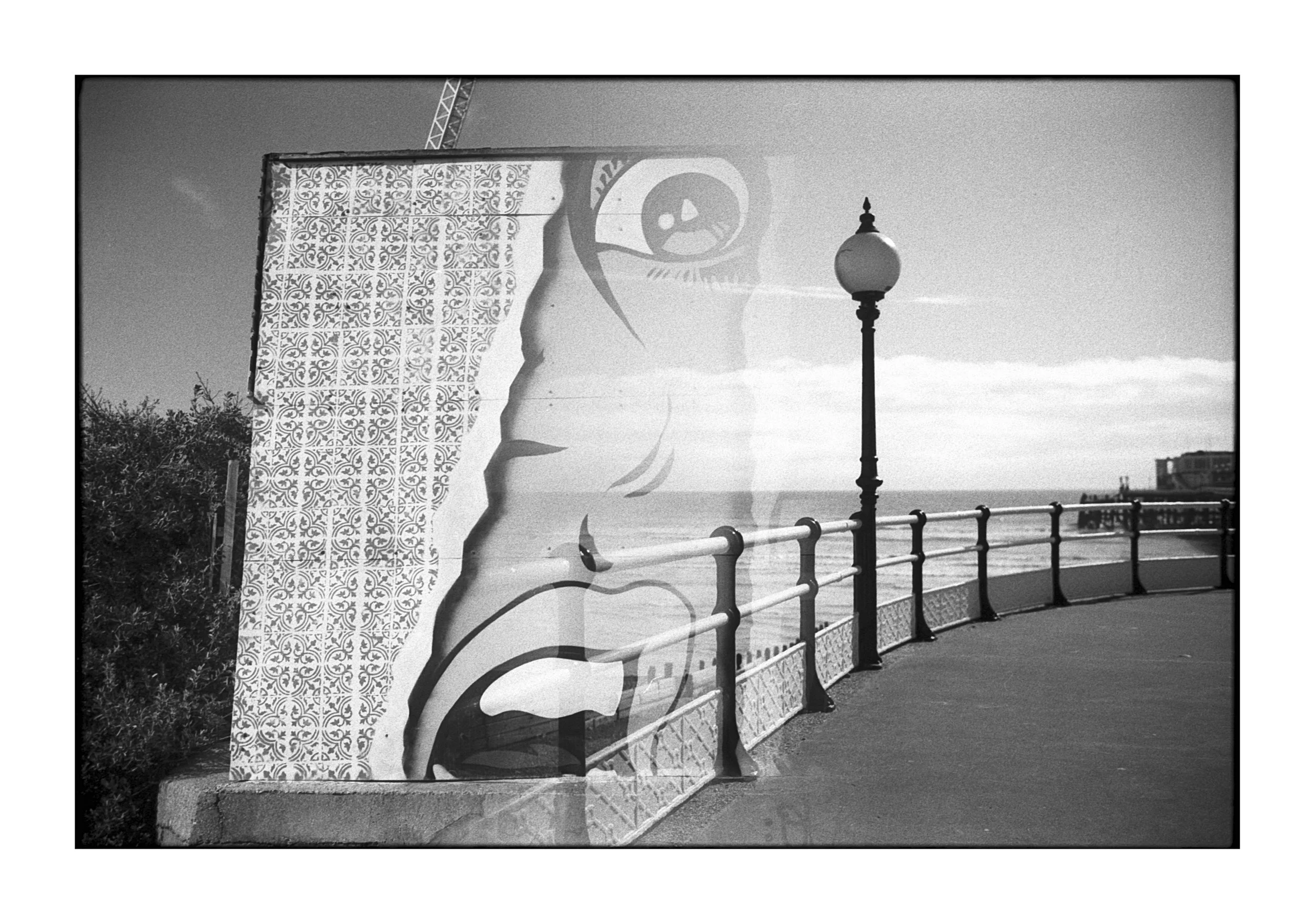 A black-and-white photograph of a coastal promenade with a decorative mural on a wall, a street lamp, and an ocean view in the background.