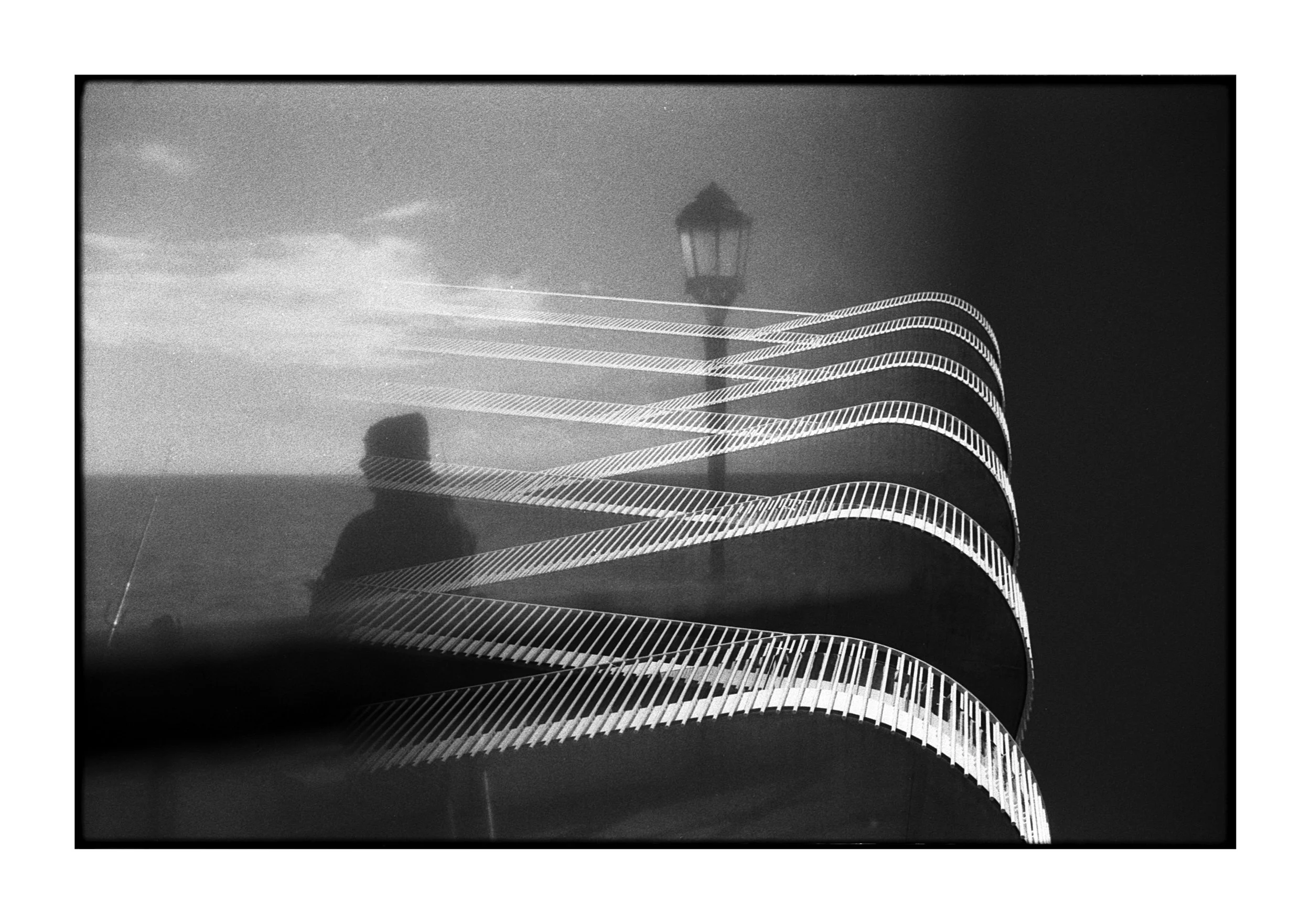 Black and white photo of a spiral staircase with a person reflection, a lamppost in the background, and clouds in the sky.