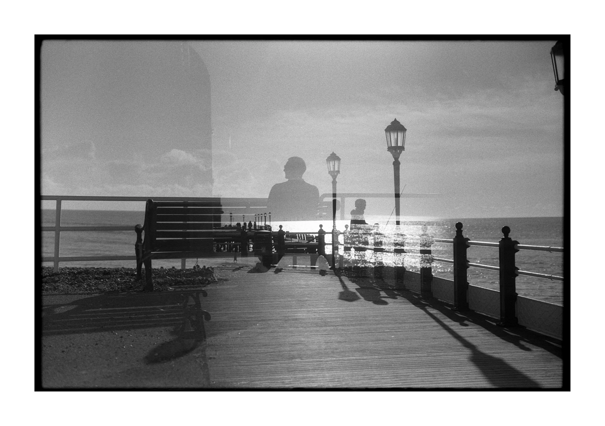 A black and white photo of a pier with benches, street lamps, and railings, with reflections of a woman's silhouette and other figures in the water, and the ocean in the background.