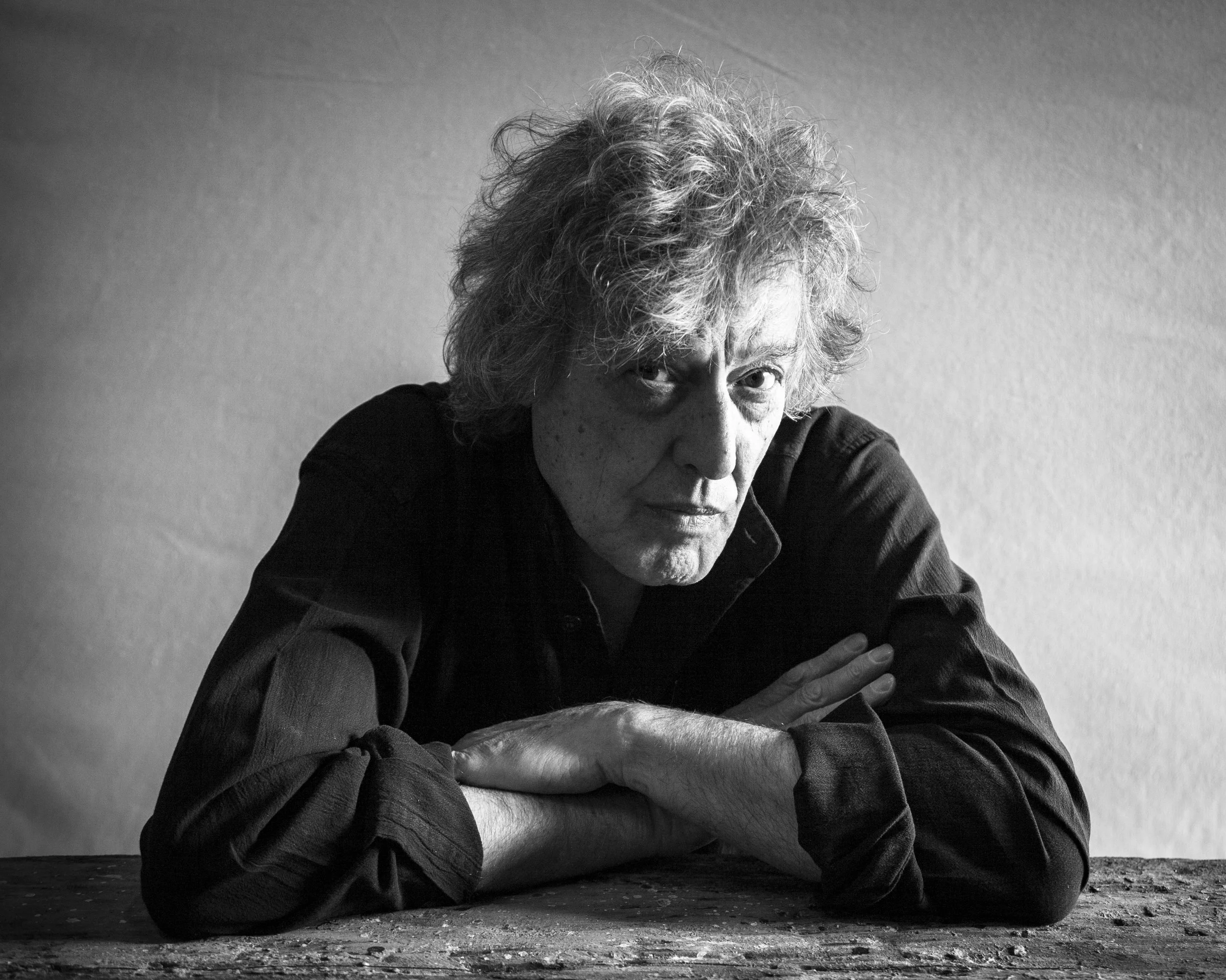 Monochrome portrait of writer Sir Tom Stoppard with natural light. He has his arms folded and is looking at the camera.