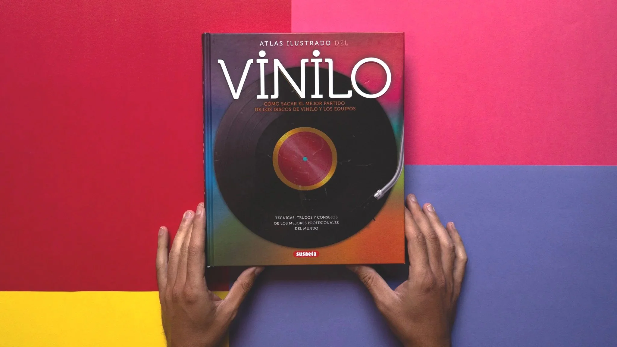 Libro titulado 'Atlas ilustrado del vinilo' con una imagen de un disco de vinilo en la portada, sostenido por dos manos sobre un fondo de papeles de colores