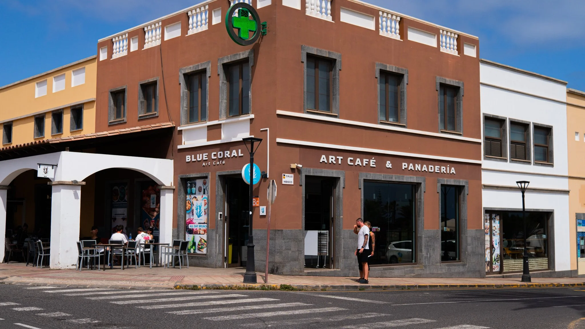 Edificio con un anuncio de farmacia y un café llamado 'Blue Coral Art Café & Panadería', con personas sentado en la acera y una persona de pie en la esquina de la calle