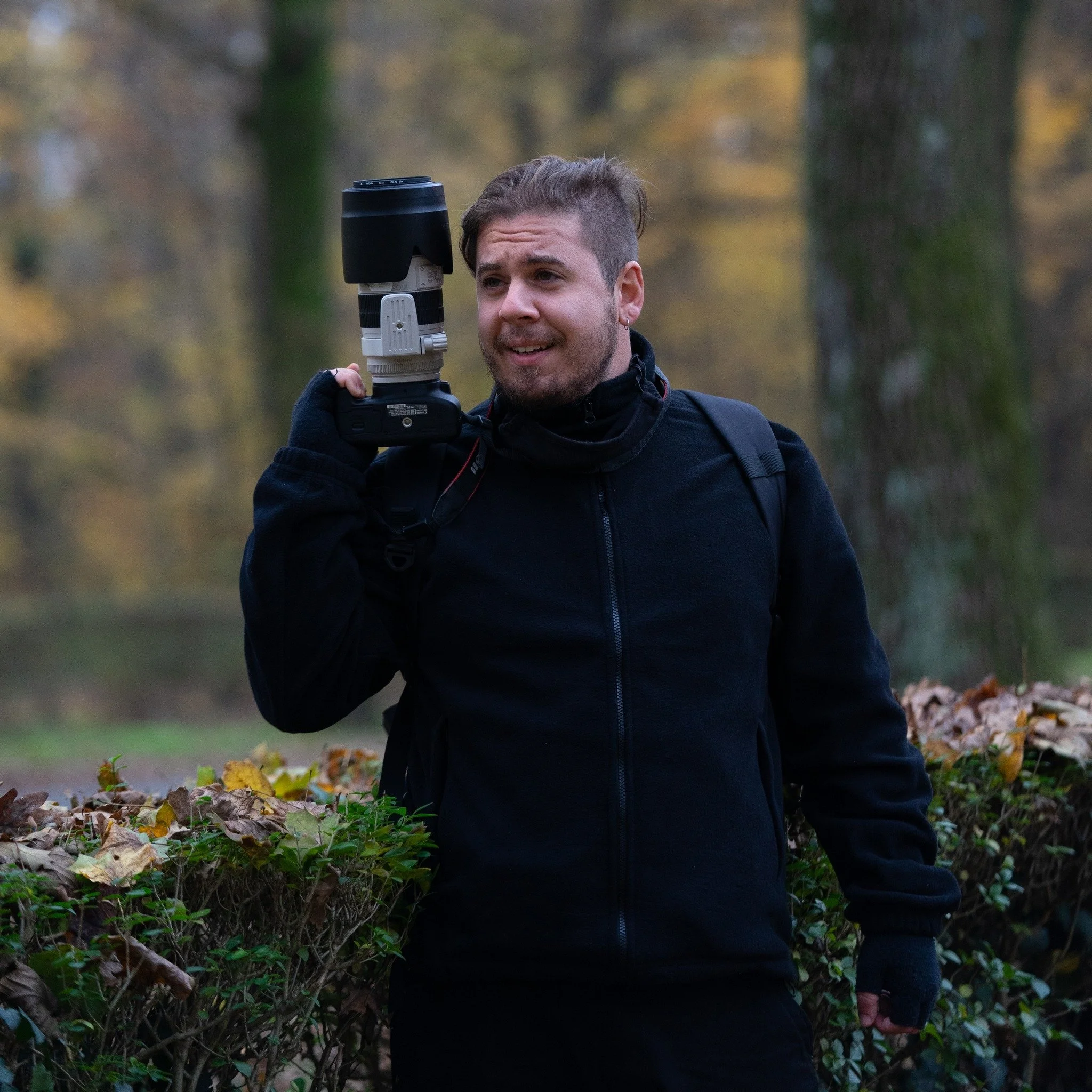 A young man in a black jacket holding a professional camera with a telephoto lens over his shoulder, standing outdoors in a wooded area with trees and fallen leaves.