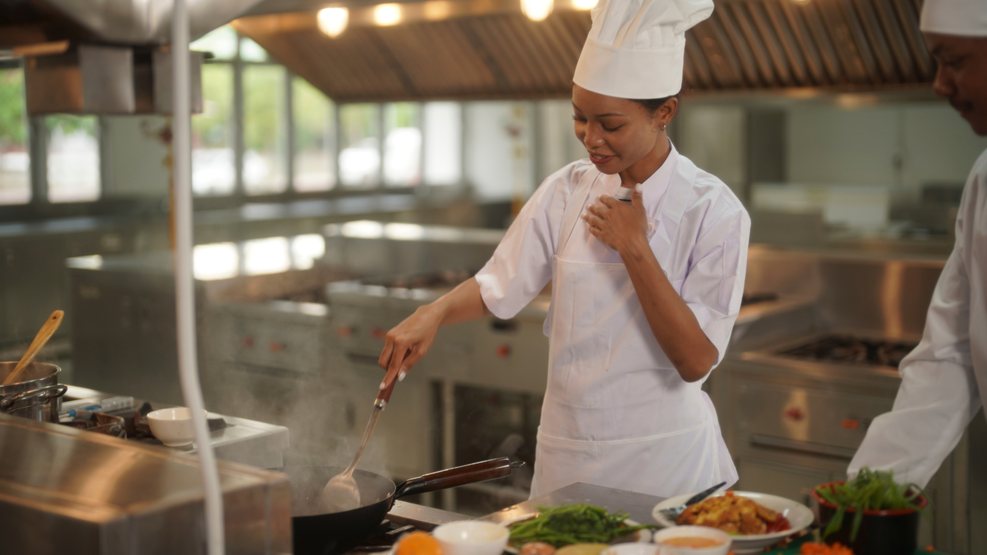 Chef com uniforme e touca cozinha numa frigideira, enquanto outro profissional prepara pratos numa cozinha profissional equipada em inox.