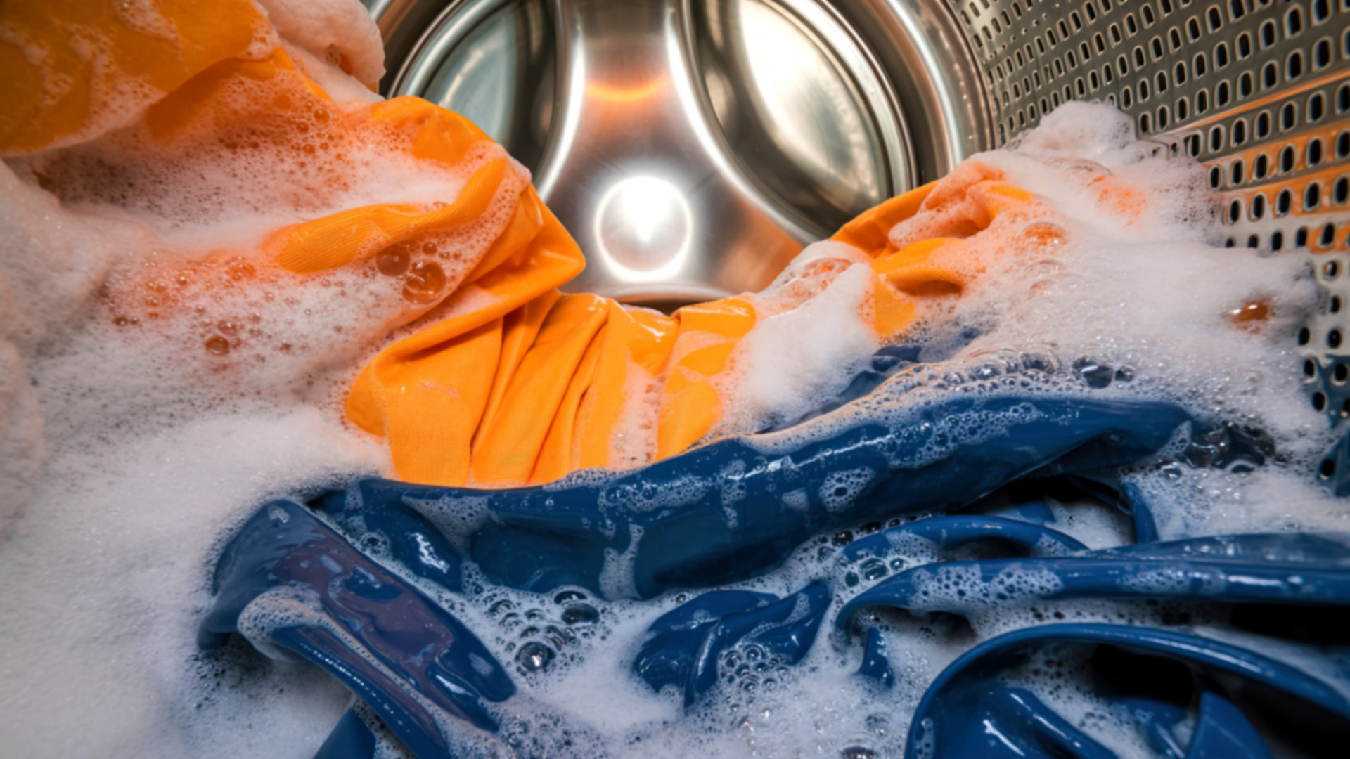 Têxteis azuis e laranja envolvidos em espuma no interior de uma máquina de lavar industrial, durante um ciclo de lavagem.